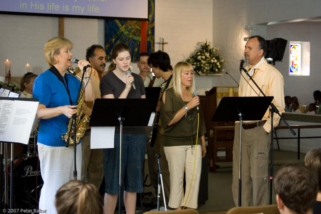 Eine Gruppe von Menschen versammelt sich in einem Raum mit einer Wandbanner, Kerzen, Papier, Mikrofonen, einem Fotorahmen und Blumen.