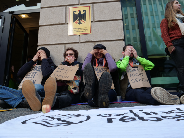Eine Gruppe von Menschen sitzt auf dem Boden und hält Schilder, mit einem Banner und einem Apfel auf der Straße, eine stehende Frau rechts und ein Gebäude mit Glasfenstern und einer Tür im Hintergrund.