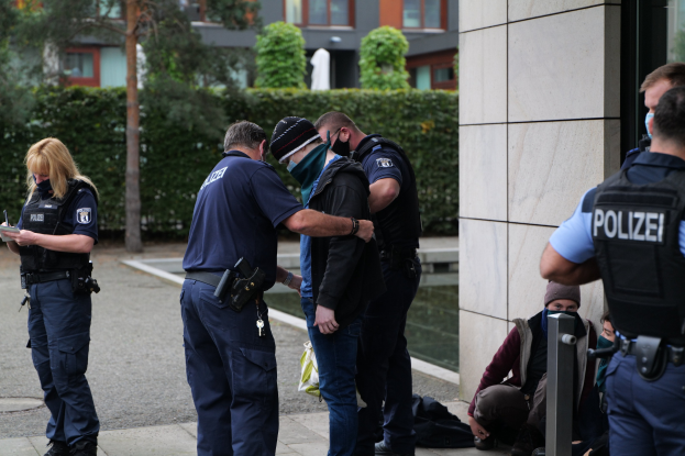 Deutsche Polizeibeamte, die in einer Gruppe vor einem Gebäude stehen, mit einer Frau, die ein Buch und einen Stift hält, und mehreren Menschen in der Nähe, umgeben von Grünflächen und städtischen Strukturen.