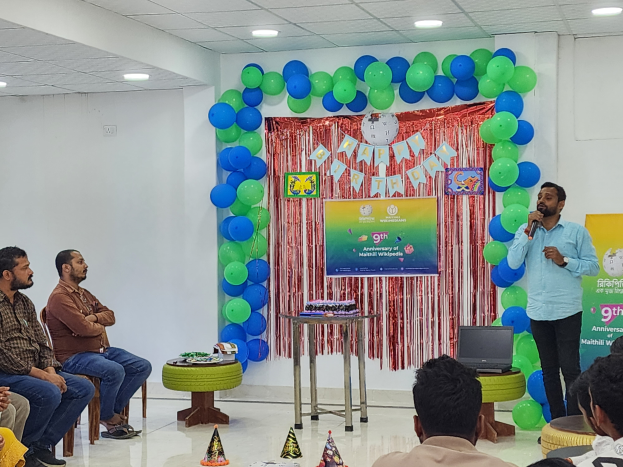 Eine Gruppe von Menschen sitzt auf Stühlen, die einer Bühne zugewandt sind, die mit Ballons, einem Banner, einer Uhr und anderen Gegenständen dekoriert ist, mit einer Person auf der Bühne, die ein Mikrofon hält, und einem Tisch mit einem Laptop, unter einer Decke mit Lichtern für eine 9. Geburtstagsfeier.