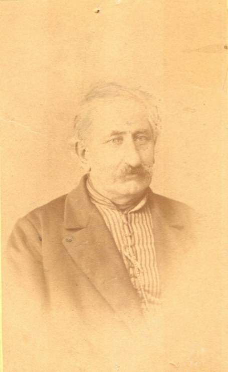 Ein Schwarz-Weiß-Porträt eines älteren Mannes mit Schnurrbart, der einen Mantel trägt und ein starkes Kinn, eine prominente Nase, dunkle Augen und einen nachdenklichen Ausdruck mit leicht geöffneten Lippen hat.