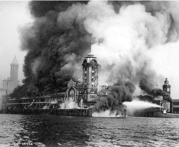 Schwarzes und weißes Foto eines großen Gebäudes, das in Flammen steht und von dem Rauch aufsteigt, mit einem Gewässer im Vordergrund und anderen Gebäuden und dem Himmel im Hintergrund.