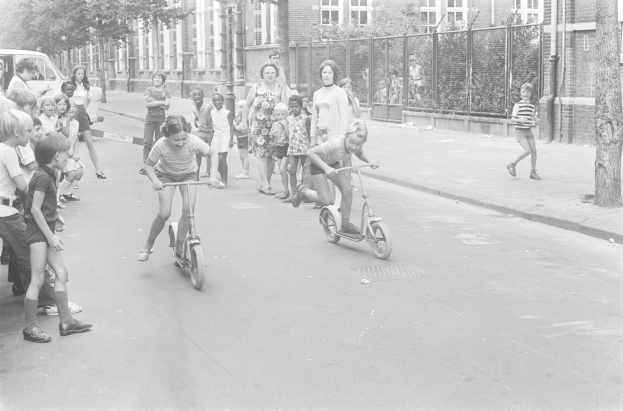 Schwarzes Bild von Kindern, die Scooter die Straße entlangfahren, mit einem geparkten Auto auf der linken Seite, Gebäuden, Bäumen, Pfählen und einem Zaun im Hintergrund.