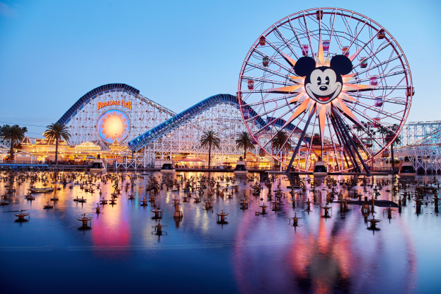 Eine lebendige Dämmerungsszene im Disneyland California Adventure mit einem riesigen Mickey-Maus-Rad im Vordergrund, einer Achterbahn im Hintergrund und Booten auf dem Wasser, mit einem farbenfrohen Himmel und Silhouetten von Bäumen.