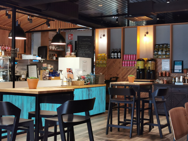 Innenraum eines Restaurants mit Holztischen, Stühlen, einem Topf mit einer Pflanze, einer Schiefertafel, einem Kühlschrank, einem Feuerlöscher, Regalen mit Flaschen, Schränken mit Gegenständen, einem gerahmten Foto an der Wand und Deckenleuchten.