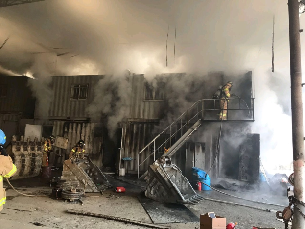Feuerwehrleute in Helmen verwenden Schläuche, um ein Lagerfeuer zu löschen, mit Rauch, der aus dem Gebäude aufsteigt, Treppenstufen mit Geländern, die zum Eingang führen, ein Kettenpfahl, eine fensterumrandete Wand im Hintergrund und verstreute Trümmer, einschließlich Kartons und Rohre auf dem Boden.