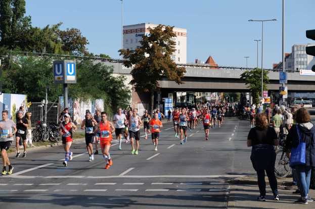 Gruppe von Menschen, die bei einem Marathon auf einer von Bäumen gesäumten Straße mit Laternen, Schildern, Fahrrädern, einem Zaun, Gras, einer Brücke, Gebäuden und einem klaren blauen Himmel laufen.
