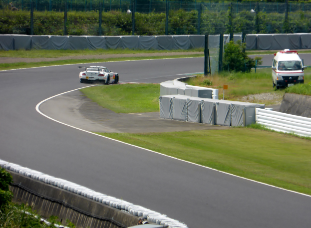 Ein Rennwagen fährt auf einer Rennstrecke mit einem Polizeiauto im Hintergrund, umgeben von Gras, Pflanzen, Bäumen und einem Zaun, mit einem Fahrzeug auf der rechten Seite und Text unten.