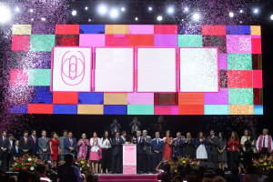 Eine Gruppe von Menschen auf einer Bühne mit einem Podium, Blumengestecken auf beiden Seiten und einem großen Bildschirm im Hintergrund während der Eröffnungszeremonie des 2018 China International Film Festival.