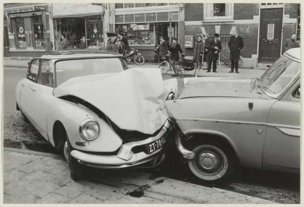 Schwarzes und weißes Foto von zwei beschädigten Autos am Straßenrand mit Menschen und Fahrrädern in der Nähe und Gebäuden im Hintergrund.