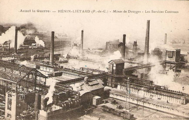 Ein altes Schwarz-Weiß-Foto einer Fabrik mit Rauch, der aus ihren Schornsteinen aufsteigt, umgeben von Gebäuden, Masten und Eisenbahnschienen, mit Text oben und unten: "Avant la Guerre - Hénin-Lietard - Mines de Douges - Les Services Centraux".