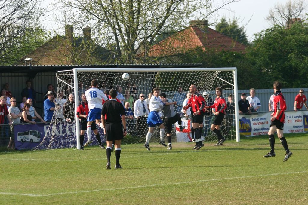 Spieler spielen Fussball auf einem Feld mit einem Tor, w├Ąhrend Zuschauer dahinter stehen, mit B├Ąumen und H├Ąusern im Hintergrund.