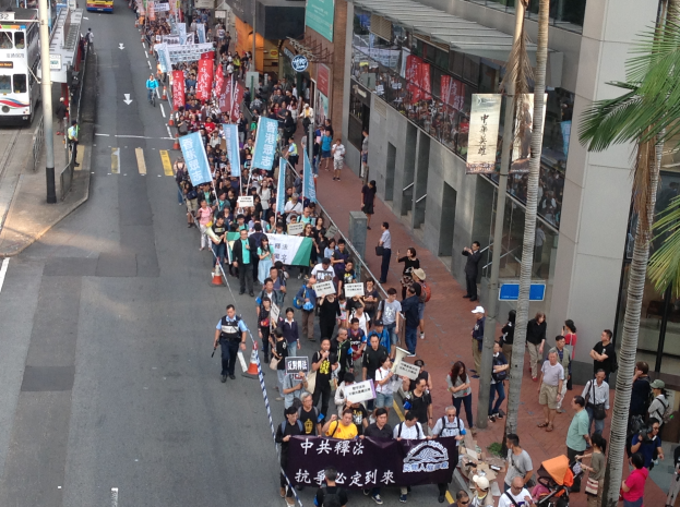 Eine große Gruppe von Menschen marschiert auf einer Straße in Hong Kong, hält Schilder und Plakate, mit Bäumen, Glasfassaden, Fahrzeugen und Schildern im Hintergrund.