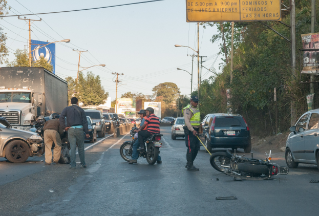Eine Gruppe von Menschen steht um ein verunglücktes Motorrad auf der Straße herum, umgeben von mehreren Fahrzeugen, darunter ein Lastwagen, und einer Hintergrundlandschaft mit Bäumen, Pfählen, Lampen und Schildern unter einem Himmel.