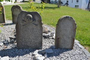 Ein Friedhof mit zwei Grabsteinen mit Totenkopf- und Knochen-Schnitzereien im Vordergrund und Gebäuden, Bäumen, Pflanzen, Gras und einer Straße im Hintergrund.