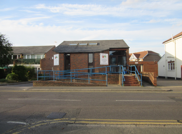 Ein Schulgebäude mit blauer Begrenzung, umgeben von Häusern, Treppen mit Begrenzungen, Texttafeln, Pflanzen, Bäumen und einem bewölkten Himmel.