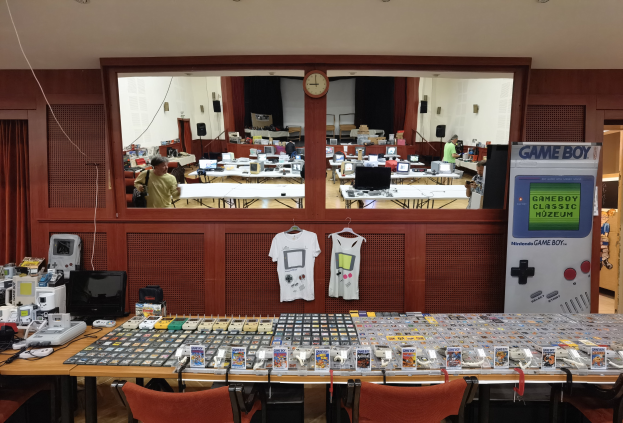 Ein Raum voller Tische und Stühle mit verschiedenen Gegenständen wie T-Shirts, Monitoren und elektronischen Geräten, mit einer Uhr an der Wand und einem Eingang zu einem anderen Raum im Hintergrund, Teil des Nintendo Game Boy Classic Museums.