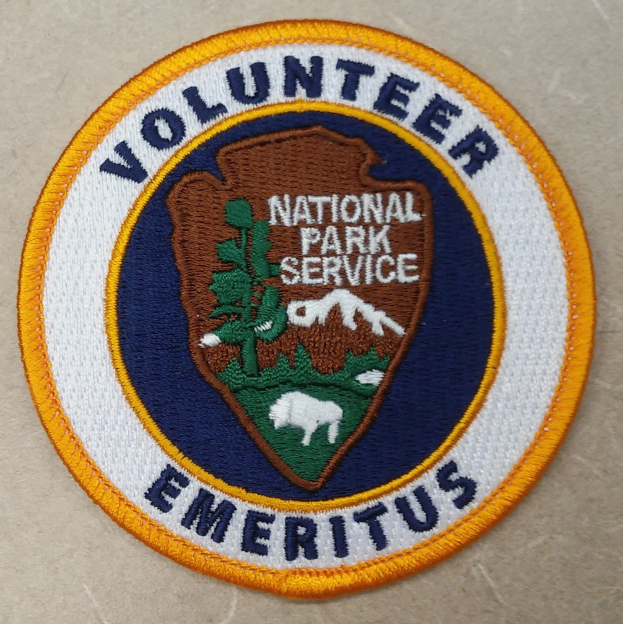 Runder Patch mit "Volunteer National Park Service Emeritus" in fetter schwarzer Schrift auf einem weißen Hintergrund, umrandet von einem blauen Rand und einem gelben Kreis.