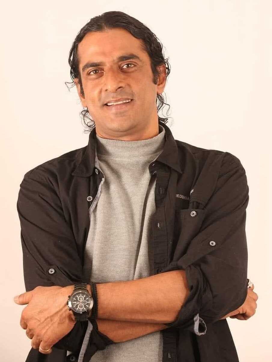 Ein Mann in einem schwarzen und aschgrauen Outfit mit überkreuzten Armen, der eine Uhr tr├Ągt und gegen einen weißen Hintergrund steht, der nachdenklich wirkt.