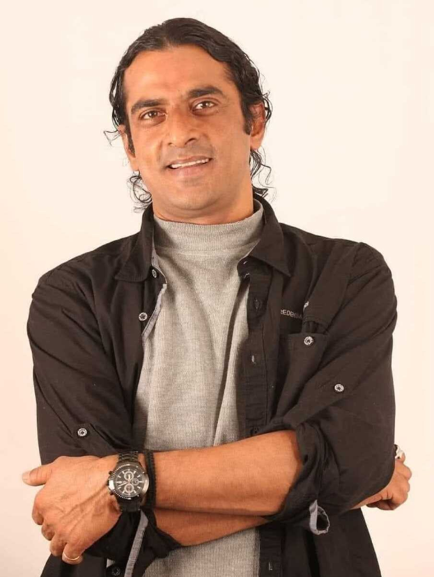 Ein Mann in einem schwarzen und aschgrauen Outfit mit überkreuzten Armen, der eine Uhr tr├Ągt und gegen einen weißen Hintergrund steht, der nachdenklich wirkt.