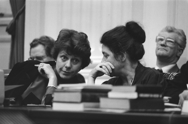 Zwei Frauen sitzen an einem Tisch in einem Gerichtssaal, wobei eine wie eine Richterin erscheint, umgeben von Büchern und Gegenst├Ąnden in einem Schwarz-Wei├č-Setting.