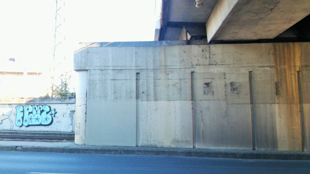 Betonwand mit Graffiti, eine Straße am Boden, ein Gebäude und ein Turm im Hintergrund und ein klarer blauer Himmel.