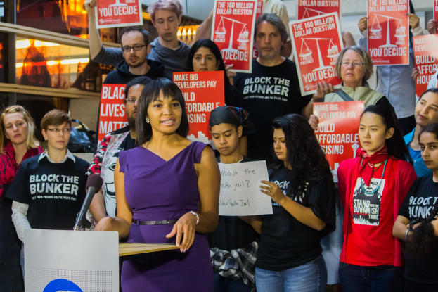 Eine Black-Lives-Matter-Aktivistin spricht an einem Rednerpult mit Mikrofon vor einer Menge mit Schildern, mit Lichtern und anderen Gegenständen im Hintergrund.