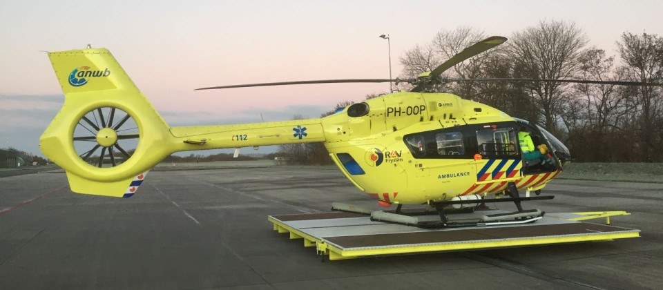Gelber Hubschrauber auf einem Flughafenvorfeld mit einer Person darin, Bäume und ein Laternenmast im Hintergrund unter einem klaren blauen Himmel.
