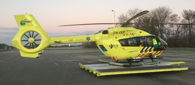 Gelber Hubschrauber auf einem Flughafenvorfeld mit einer Person darin, Bäume und ein Laternenmast im Hintergrund unter einem klaren blauen Himmel.