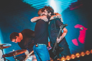 Zwei Männer umarmen sich auf der Bühne eines Konzerts, wobei einer eine Gitarre und ein anderer ein Schlagzeug spielt, unter Bühnenbeleuchtung.