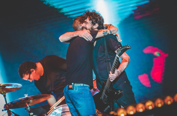 Zwei Männer umarmen sich auf der Bühne eines Konzerts, wobei einer eine Gitarre und ein anderer ein Schlagzeug spielt, unter Bühnenbeleuchtung.