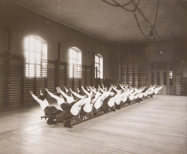 Gruppe von Menschen, die Pilates-Übungen in einem Fitnessstudio mit einer Uhr und Fenstern an der Wand durchführen.