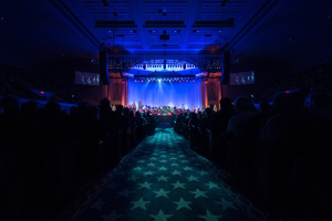 Ein großer Saal mit Sitzplätzen, eine Bühne mit Musikern und Überkopfleuchten und -fernseher, was auf ein 2019 Nationales Parteitag-Konzertsaal hindeutet.