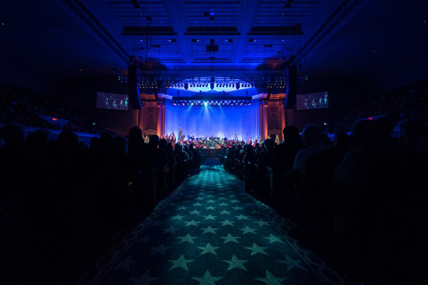 Ein großer Saal mit Sitzplätzen, eine Bühne mit Musikern und Überkopfleuchten und -fernseher, was auf ein 2019 Nationales Parteitag-Konzertsaal hindeutet.