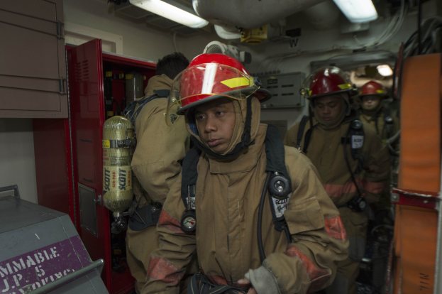 Feuerwehrleute in Uniformen und Helmen, einer hält einen Feuerlöscher, mit einem metallischen Objekt links und Hintergrundelementen einschließlich Lichter und Rohre.