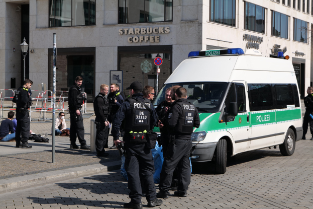 Eine Gruppe von Polizeibeamten steht vor einem Starbucks-Café mit einem Van auf der rechten Seite und einigen Menschen auf der linken Seite, während im Hintergrund ein Gebäude mit Fenstern, ein Schild, ein Laternenpfahl und ein Zaun zu sehen ist.