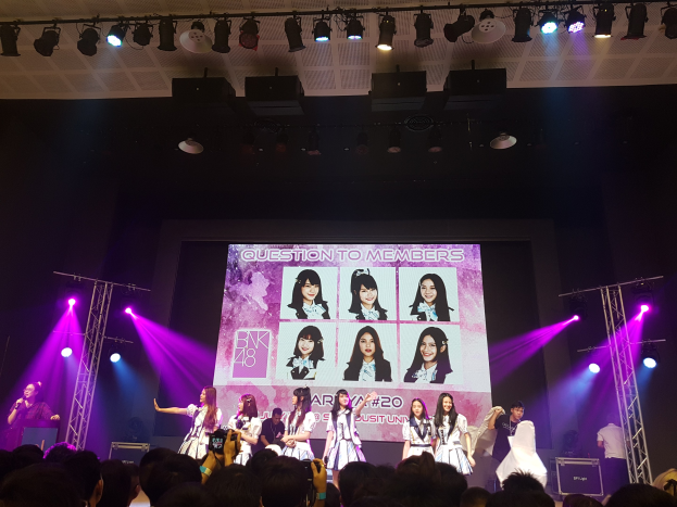 Eine Gruppe von Menschen performs auf einer hell erleuchteten Bühne mit Trägern, während ein Sänger ein Mikrofon hält und ein anderer ein Instrument spielt, während das Publikum darunter das Konzert aufzeichnet; ein großer Bildschirm im Hintergrund zeigt Bilder der K-Pop-Gruppe BK48.