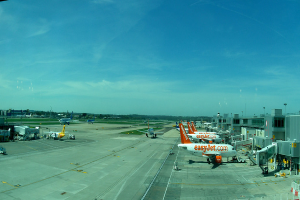 Eine Luftaufnahme eines Flughafens mit Flugzeugen, GebÃ¤uden, Masten, Fahrzeugen und BÃ¤umen, mit einer StraÃe unten und einem Zaun im Vordergrund.