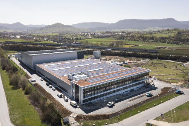 Luftaufnahme eines großeen Gebäudes mit Solarpanelen auf dem Dach, umgeben von Fahrzeugen, Bäumen, Gras und Strommasten, mit Hühgeln und einem klaren blauen Himmel im Hintergrund.