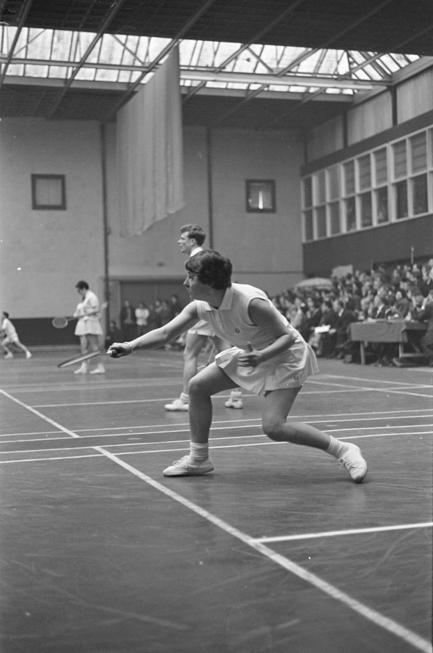 Schwarz-weißes Foto von zwei Frauen, die Badminton auf einem Court spielen, jede hält einen Schläger, mit Zuschauern rechts und einer fensterumrandeten Wand im Hintergrund.