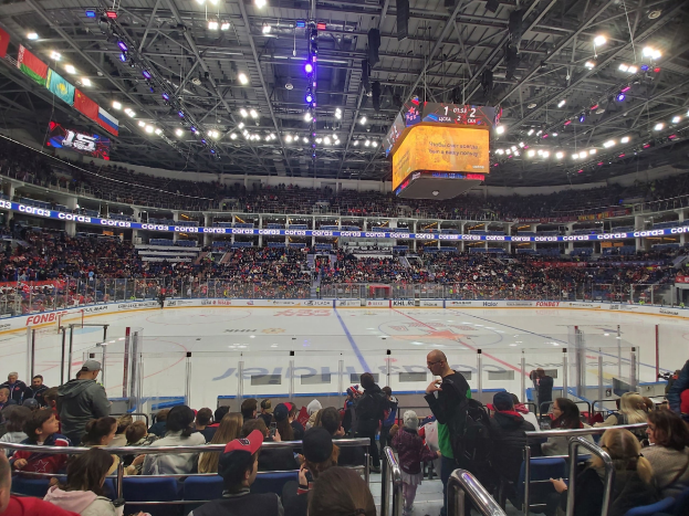 Eine große Menschenmenge schaut einem Hockey-Spiel in einer hell erleuchteten Arena zu, mit Zuschauern, die auf und um die Eisfläche sitzen und stehen, die von einem Zaun umgeben ist, und Bannern, die von der Decke hängen.