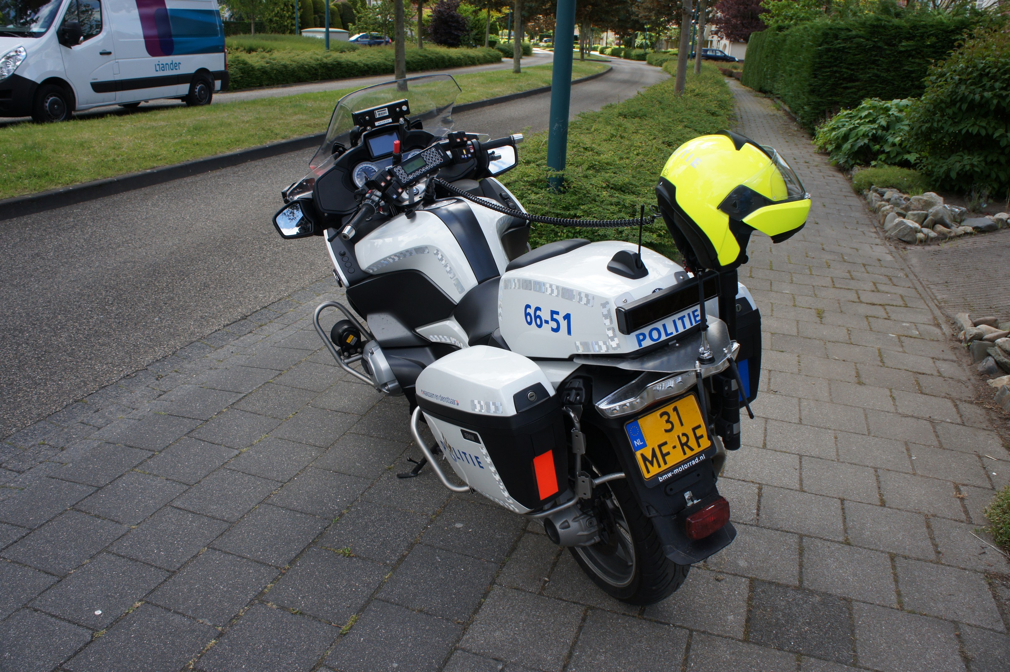 Ein Polizei-Motorrad am Rande der Straße mit einem Helm darauf, umgeben von Bäumen, Masten, Gebäuden, Pflanzen, Gras, Steinen und anderen Fahrzeugen.