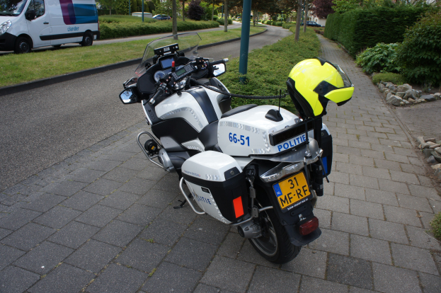 Ein Polizei-Motorrad am Rande der Straße mit einem Helm darauf, umgeben von Bäumen, Masten, Gebäuden, Pflanzen, Gras, Steinen und anderen Fahrzeugen.