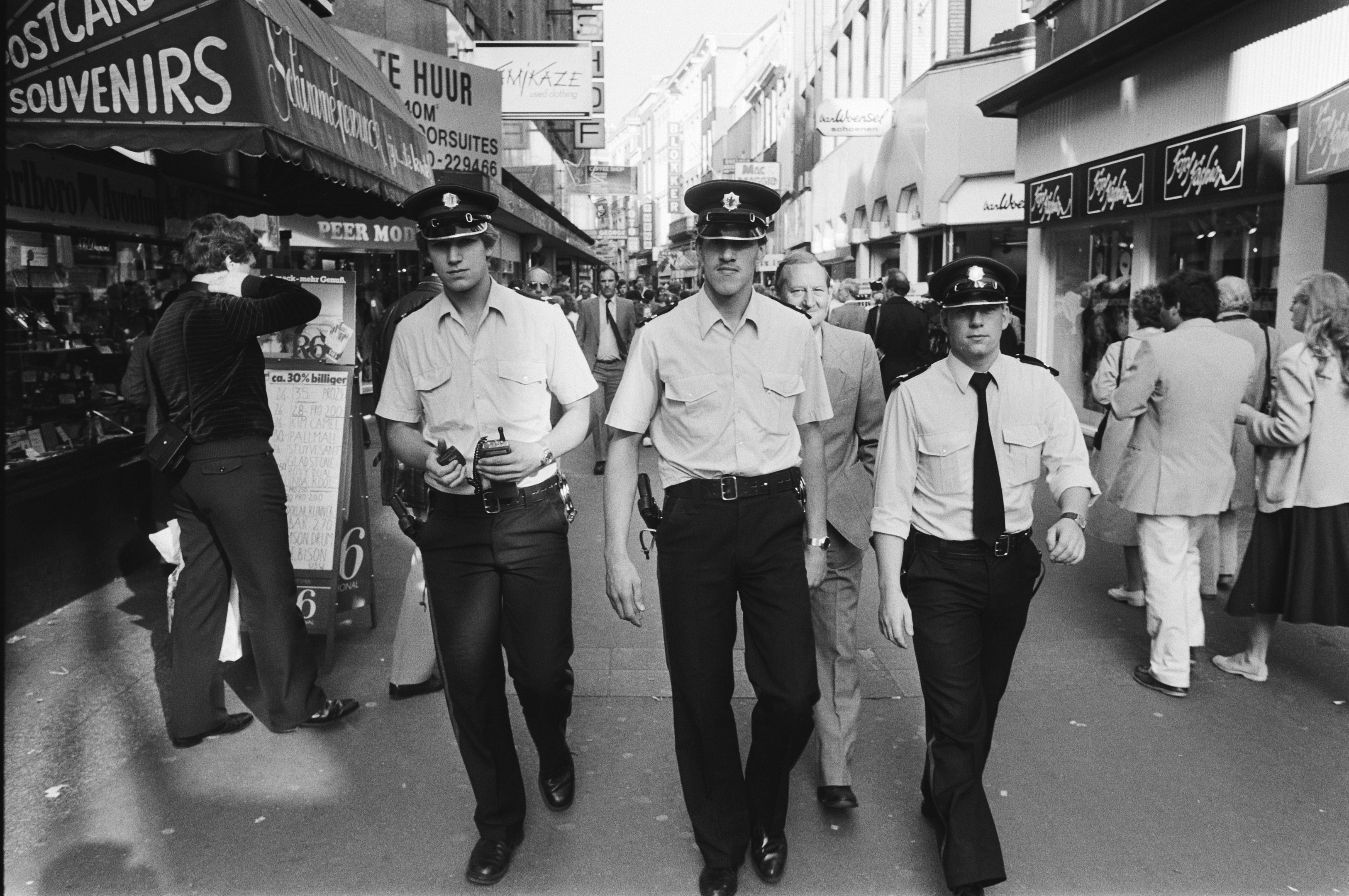 Drei Polizisten in Uniform gehen eine belebte Stadtstraße entlang, die von hohen Gebäuden und Geschäften gesäumt ist, mit Passanten und einem sichtbaren Himmel im Hintergrund.