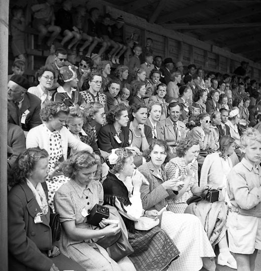 Schwarzes und weißes Foto einer Menge, die in einem Stadion mit einer hölzernen Wand und einem Dach sitzt, wobei einige Kameras halten.