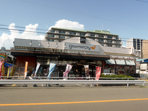 Eine belebte Stadtstraße mit Gebäuden, Fahrzeugen, Straßenmöblierung, Grünflächen und einem Restaurant, das Speisen in seinem Schaufenster präsentiert.