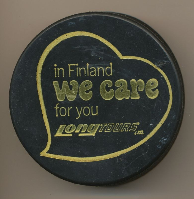 Schwarzer Eishockeypuck mit weißer Schrift "In Finland We Care For You Long Tours" auf cremefarbenem Hintergrund.