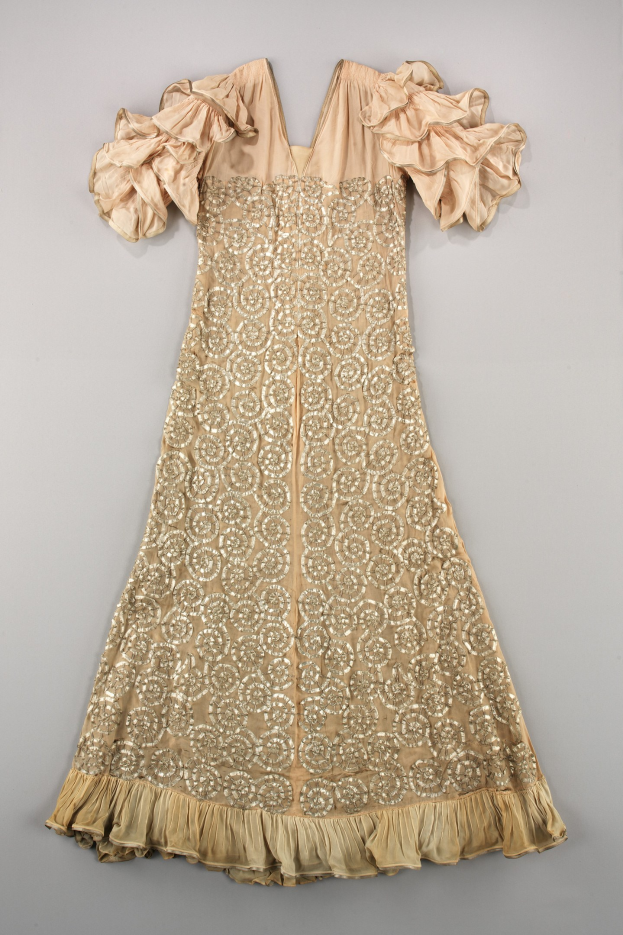 Ein cremefarbenes Kleid mit goldener Stickerei und Rüschenärmeln, das auf einer weißen Oberfläche präsentiert wird und mit aufwändigen Designs und Mustern verziert ist.