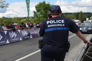 Polizist in Uniform mit Mütze und Funkgerät vor einer Menge mit Schildern, Fahrzeugen, Bäumen, Gebäuden und einem bewölkten Himmel im Hintergrund.