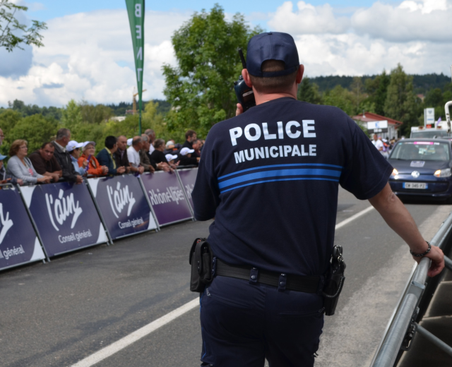 Polizist in Uniform mit Mütze und Funkgerät vor einer Menge mit Schildern, Fahrzeugen, Bäumen, Gebäuden und einem bewölkten Himmel im Hintergrund.
